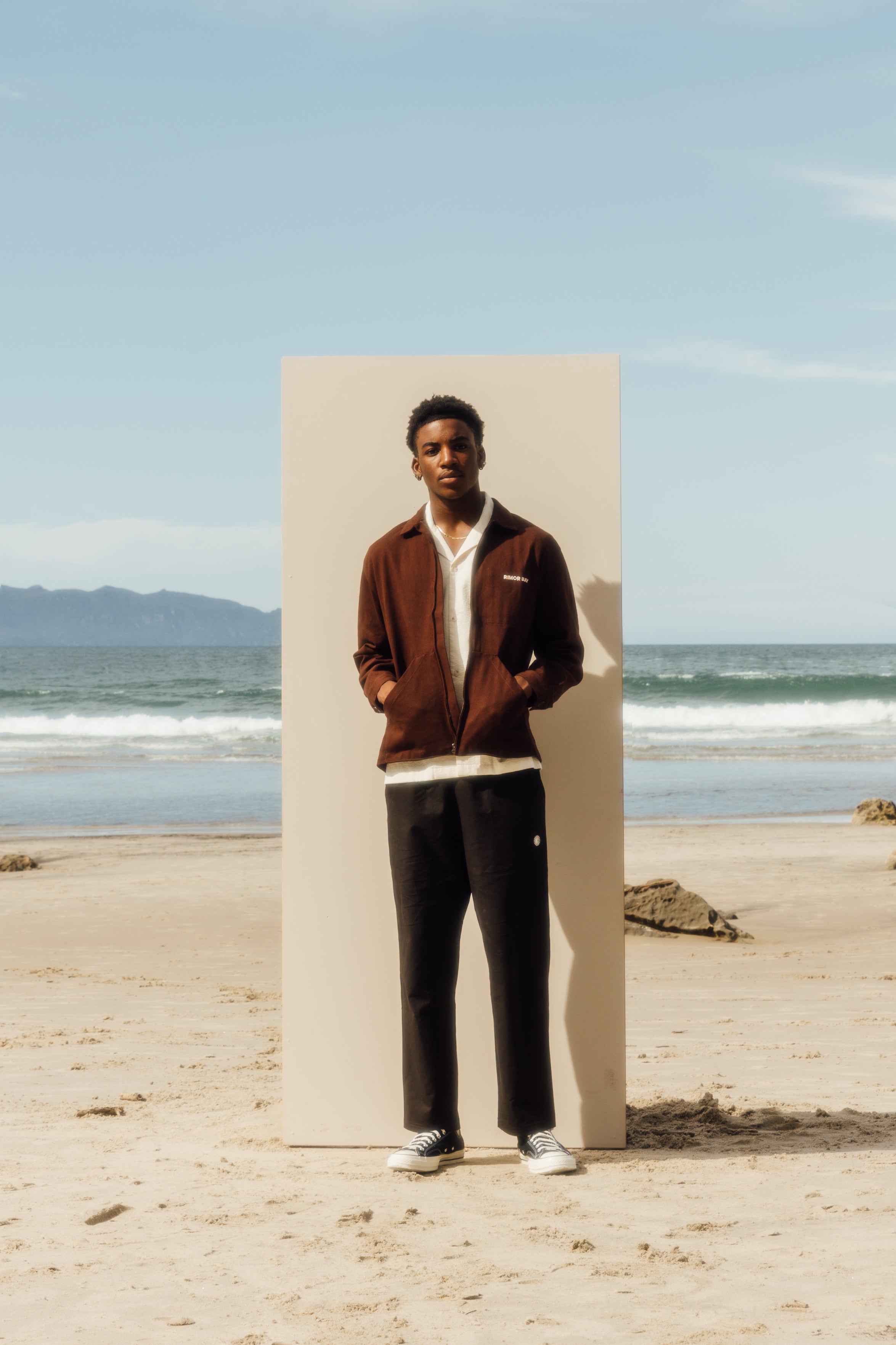 Man standing in front of a beige board at the beach, wearing a brown collared jacket, black pants, white shirt and chucks