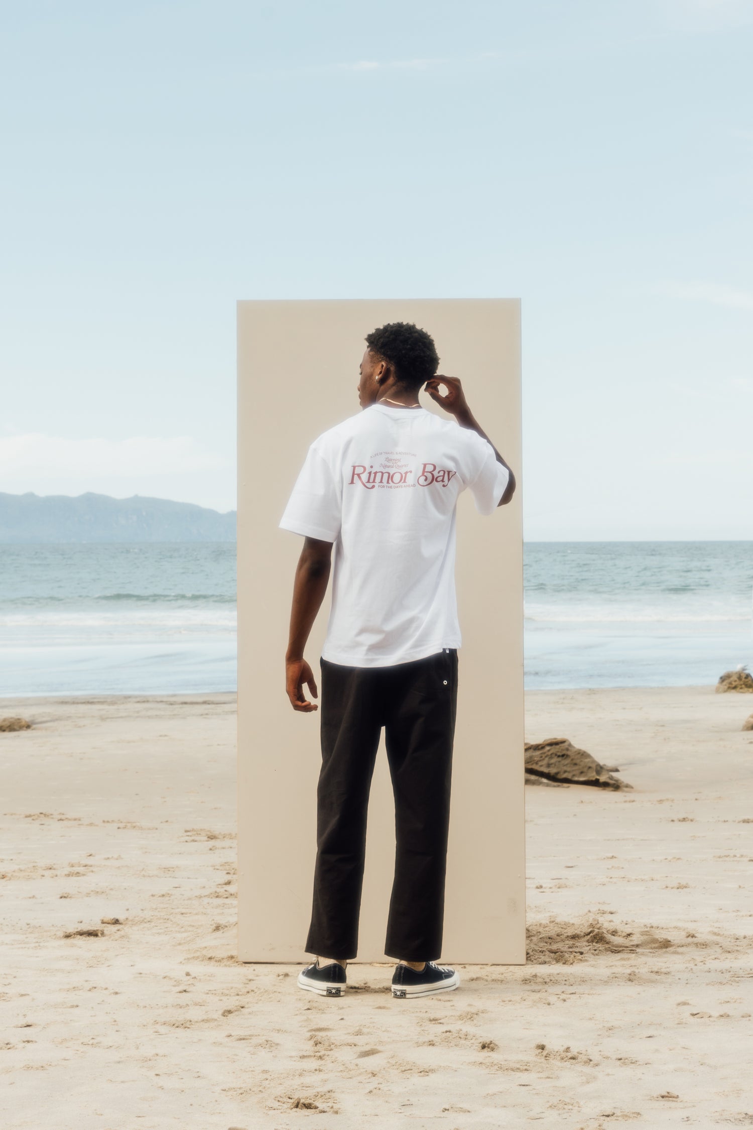 man standing on the beach in front of a beige board wearing white logo tshirt, black pants and chucks.