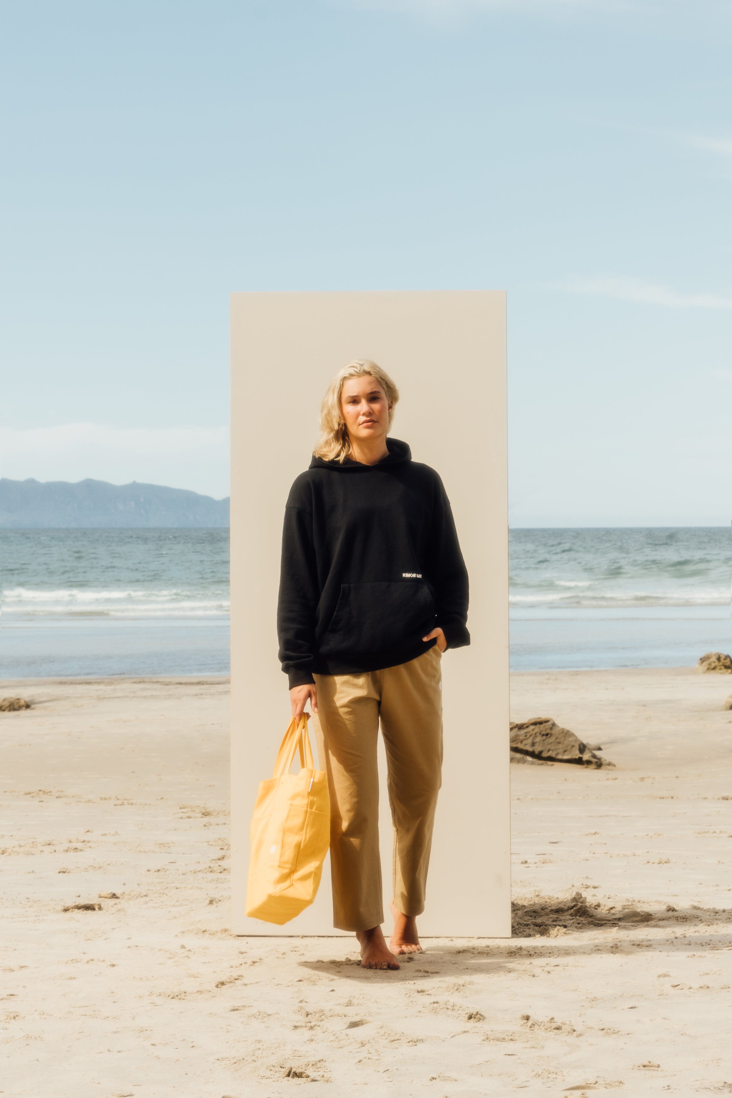 girl standing on beach in front of a beige board wearing black hoodie, beige pants, holding a yellow beach bag