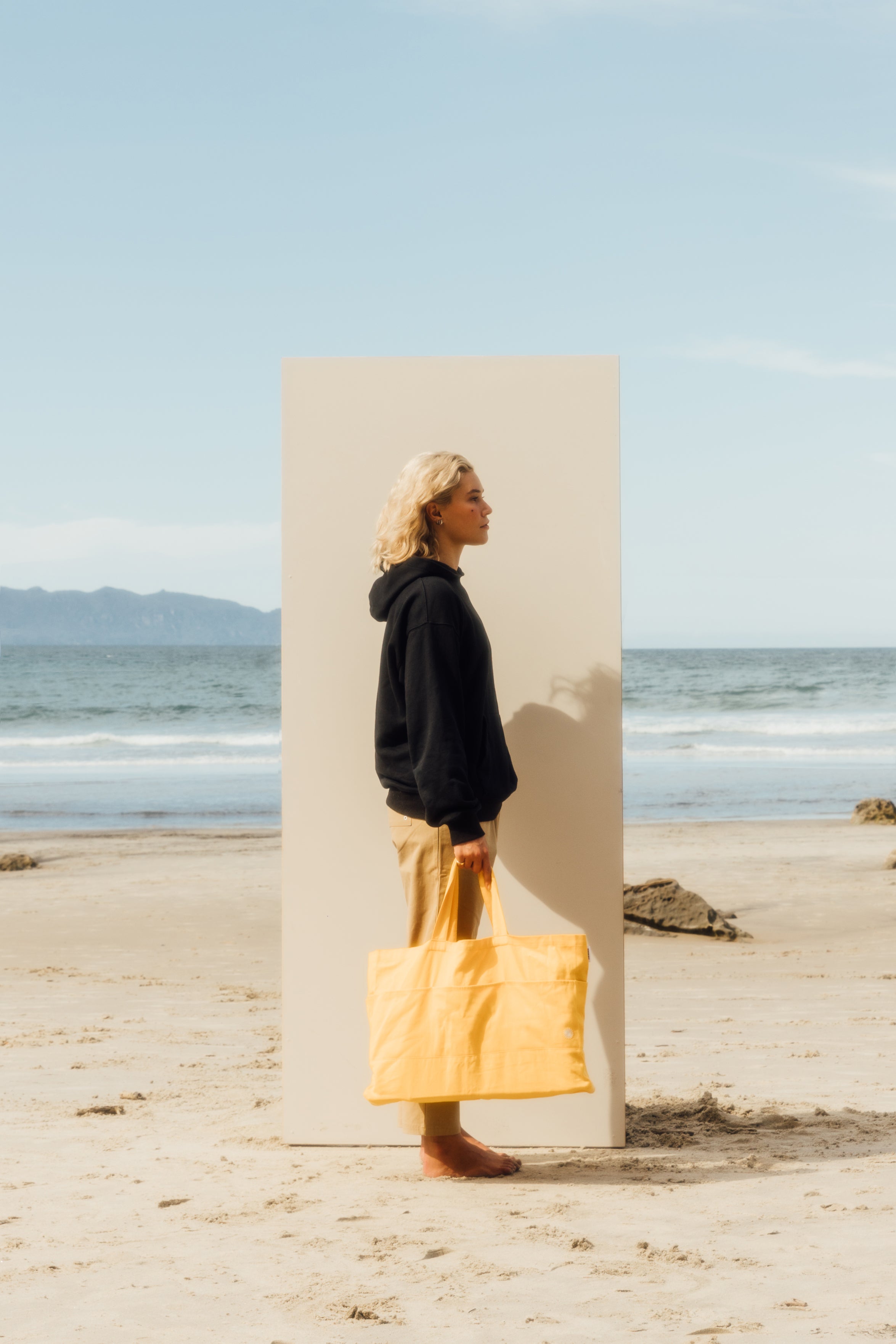 girl standing in front of beige board on the beach wearing black hoodie, beige pants, holding yellow beach bag