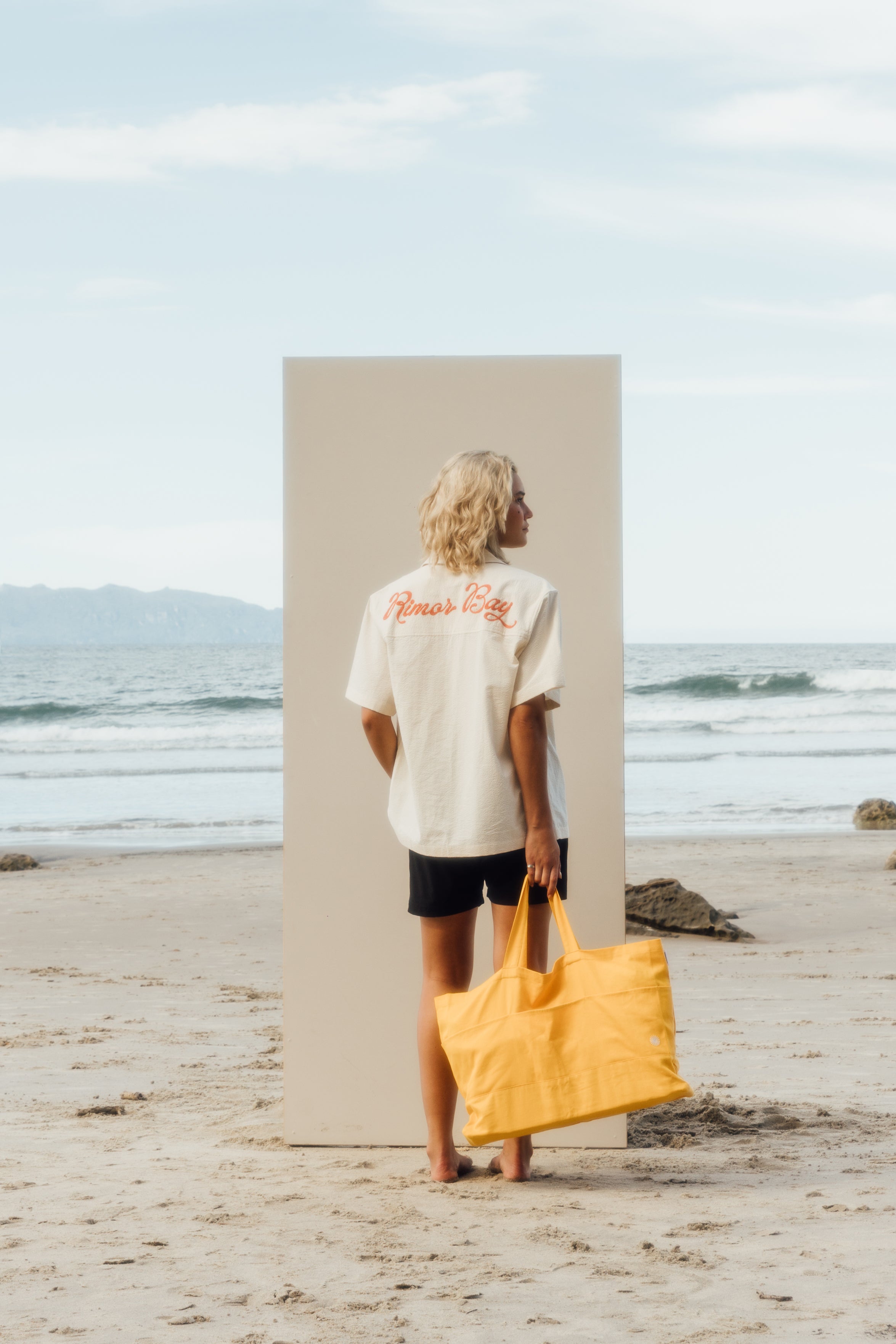 girl standing on beach in front of beige board wearing beige shirt, black shorts, holding yellow beach bag