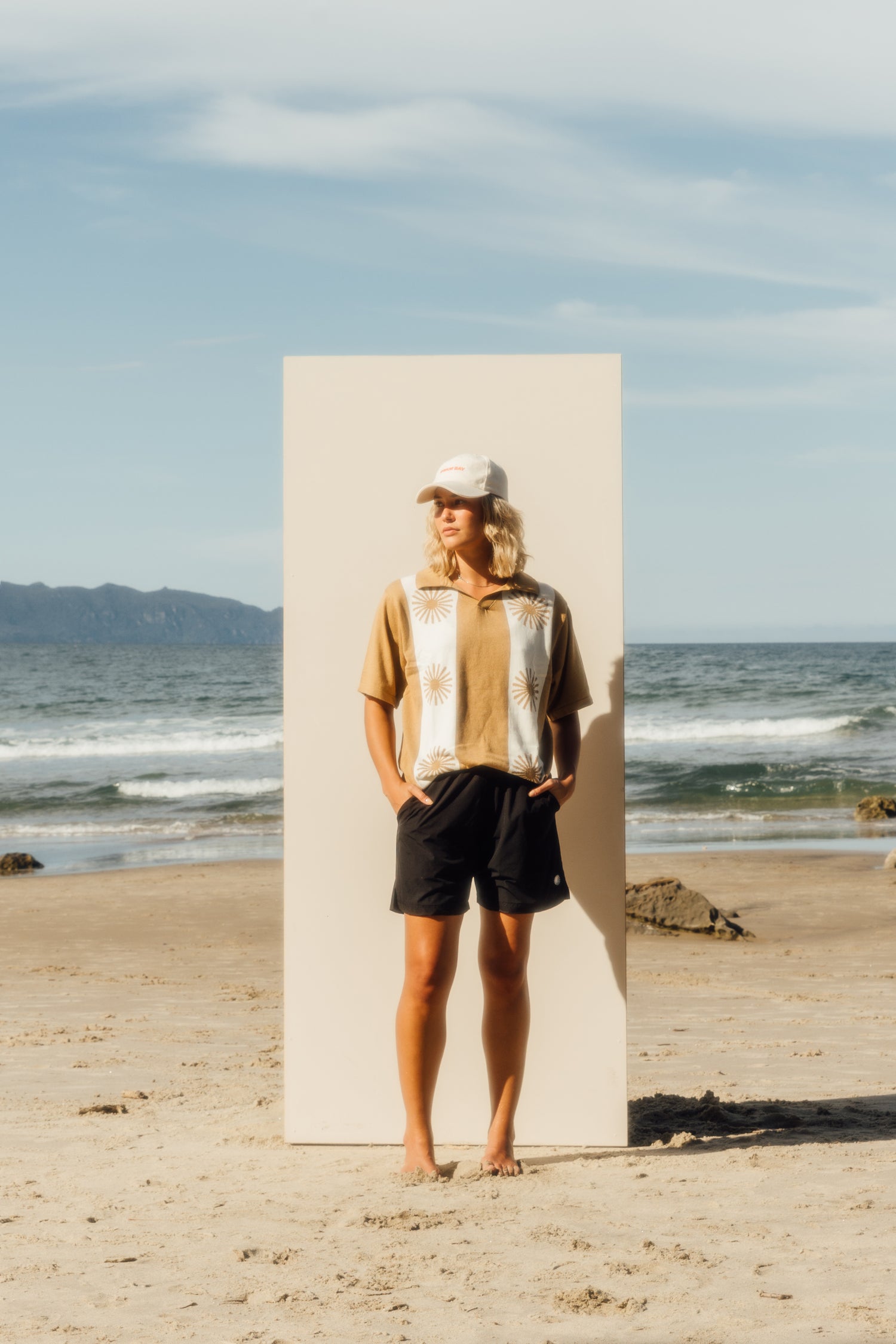 girl standing on beach in front of a beige board wearing beige hat, caramel knit top, and black shorts 