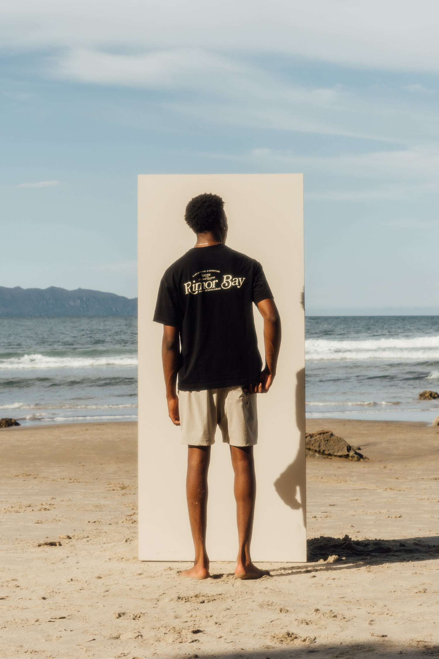 man standing on the beach in front of a beige board wearing a black logo tshirt and grey shorts