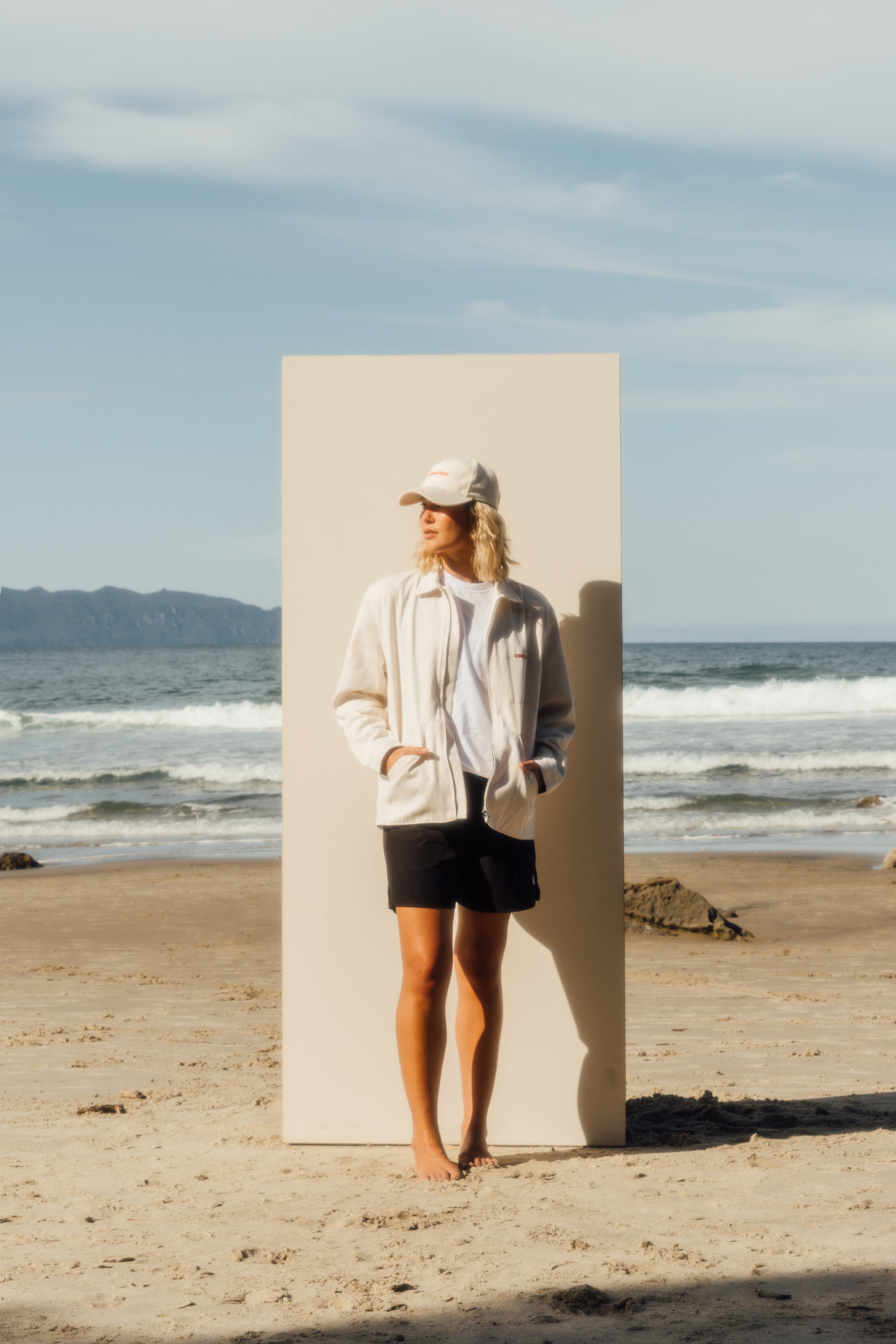 girl standing on beach in front of a beige board wearing beige hat, off white jacket, white tshirt, and black shorts