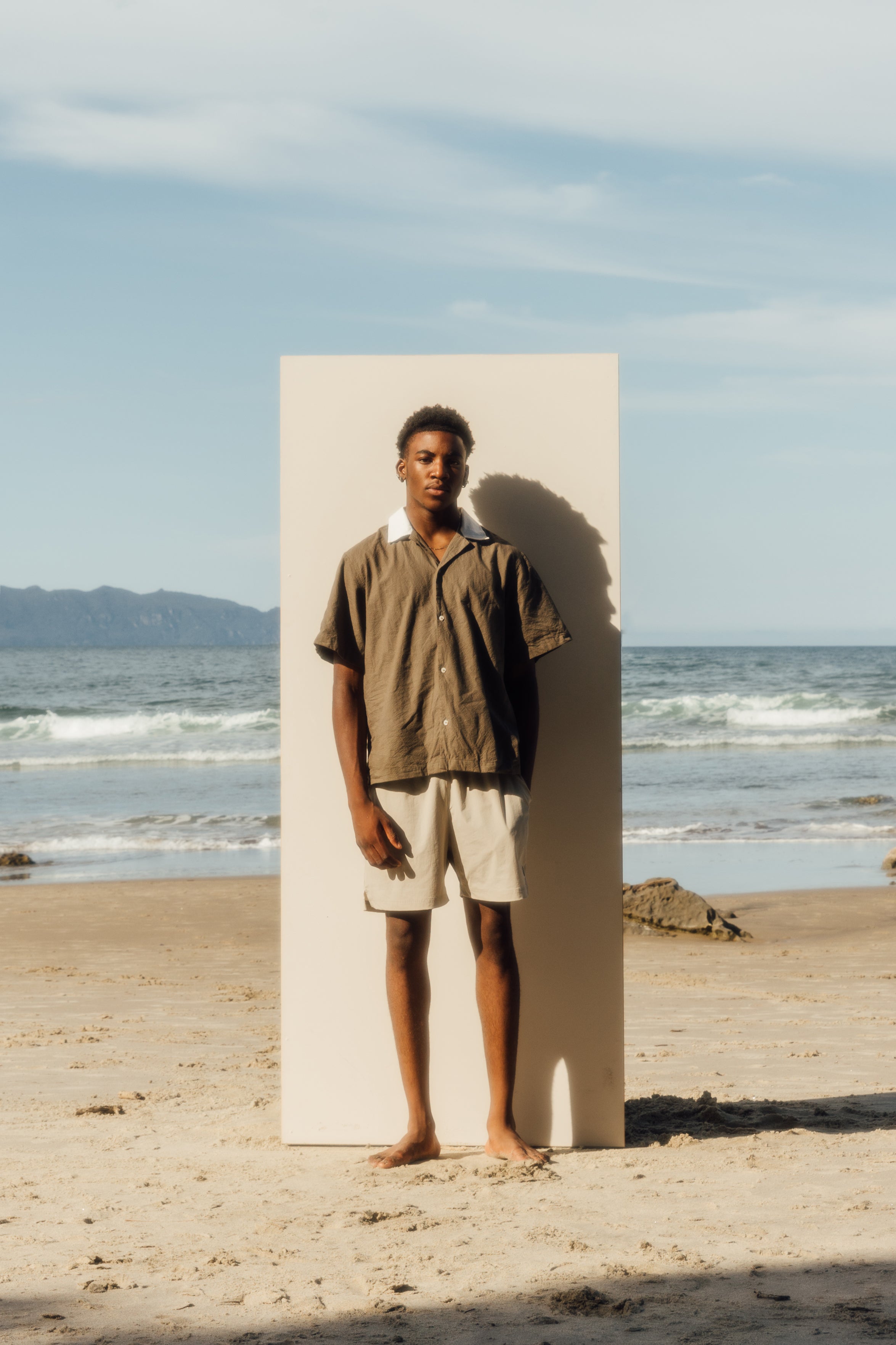 man standing on beach in front of beige board wearing khaki shirt and grey shorts