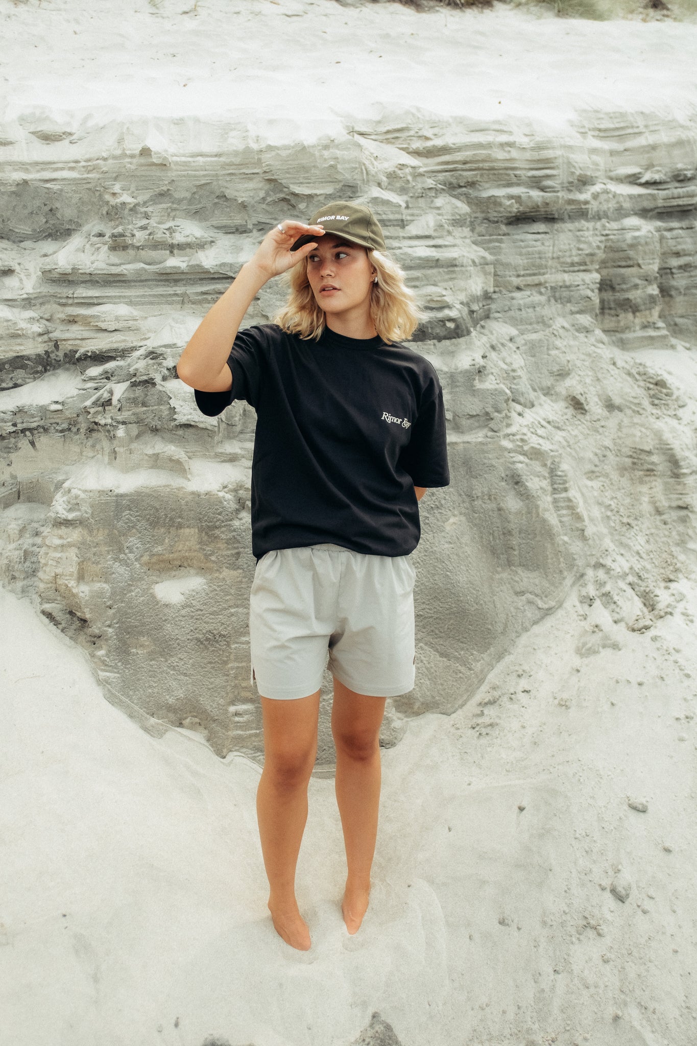 Girl wearing khaki cap with black tshirt and grey shorts standing on sand